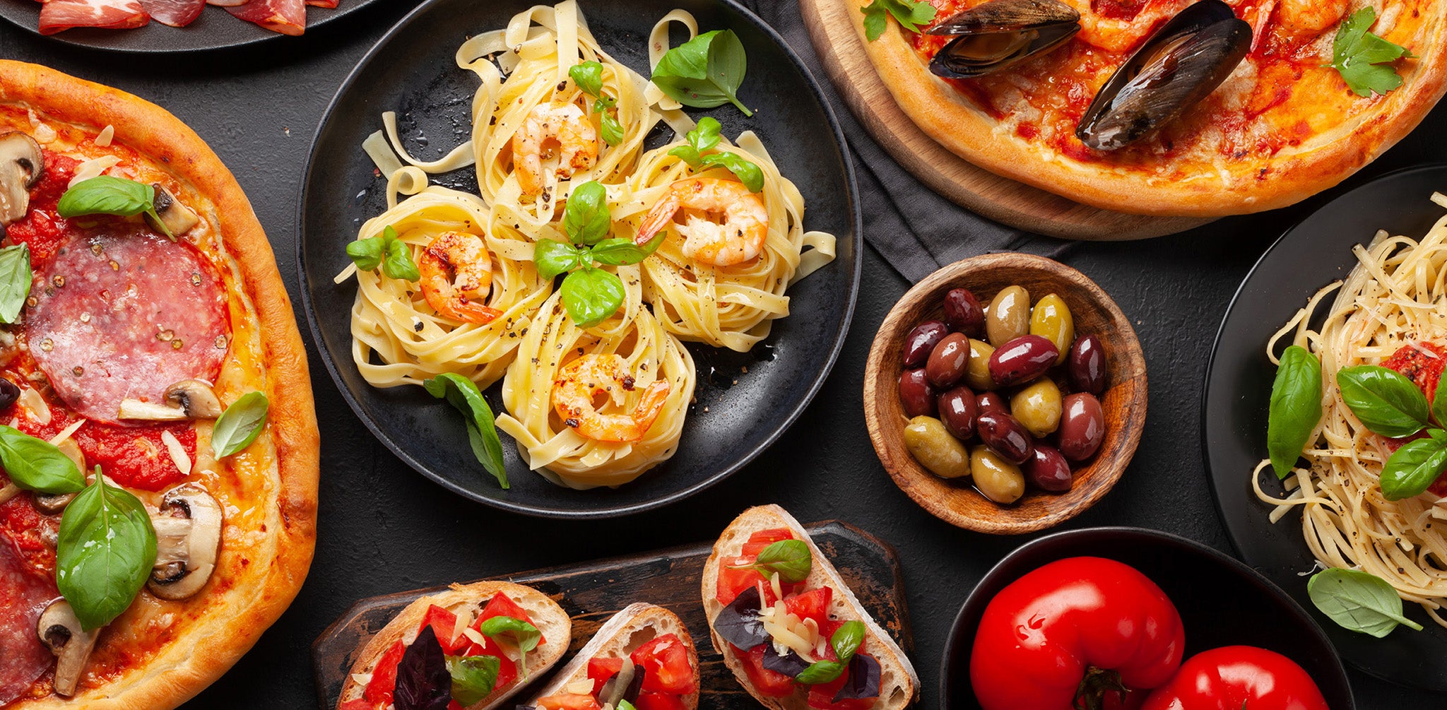 A collection of dishes: pasta nests, a tray of bruschetta, several pizzas, a plate of prosciutto, and a bowl of olives, arrange on a black table.