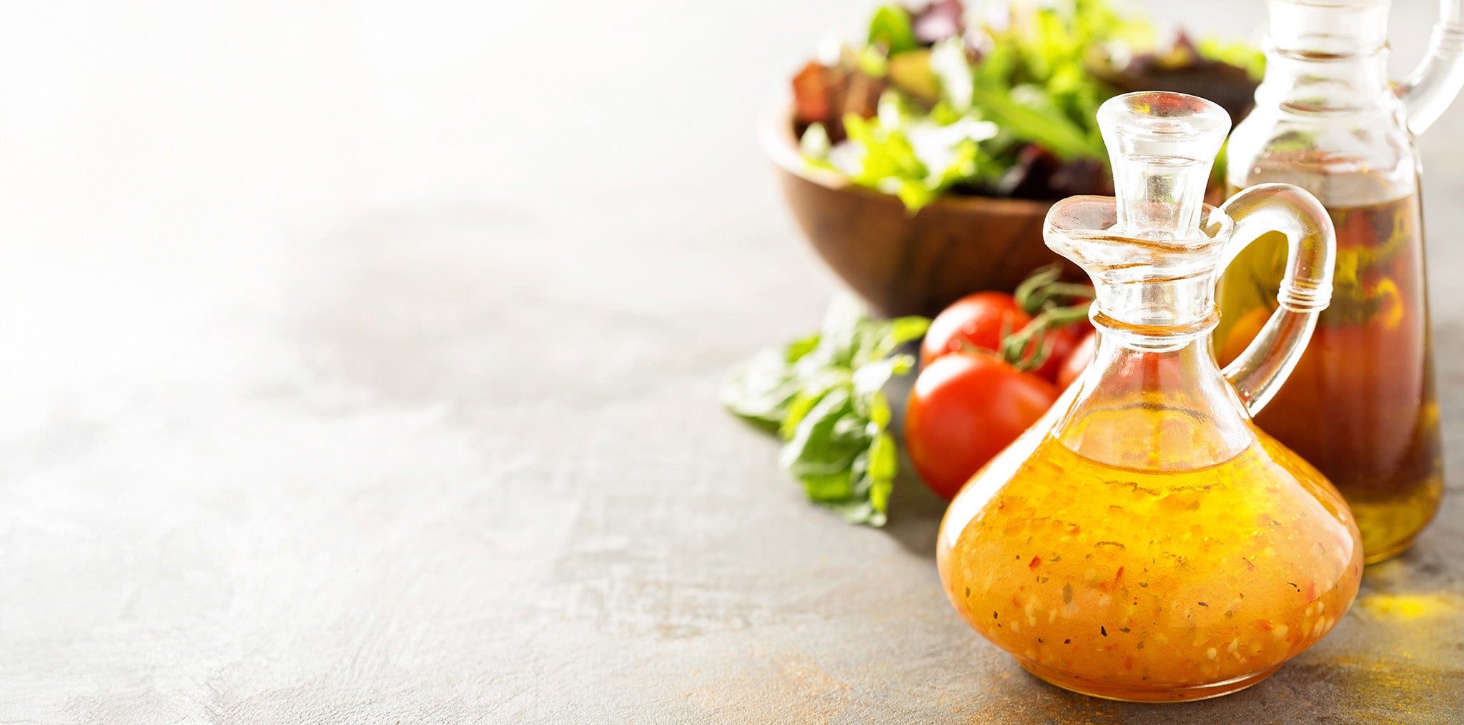 A glass carafe of viniagrette in the foreground, with tomatoes on the vine and a wooden bowl of salad in the background.
